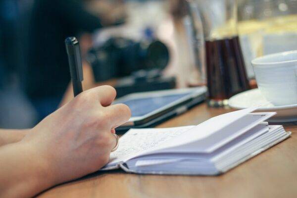 A photo of a hand taking notes with wine in background.